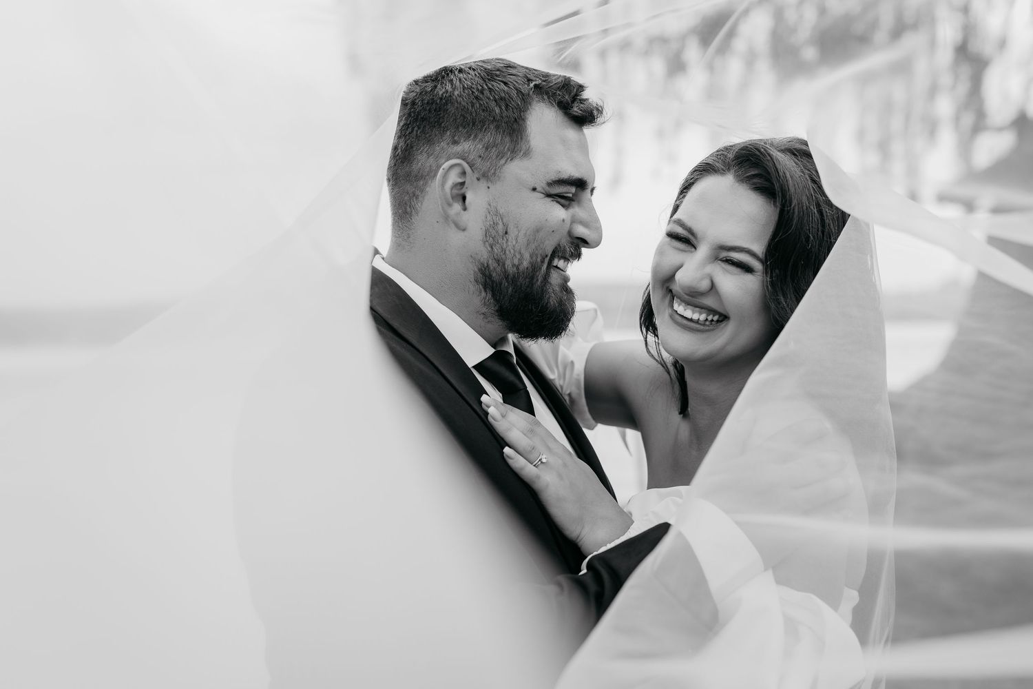 Black and white candid wedding photo showing a joyful intimate moment between newlyweds under a flowing veil.