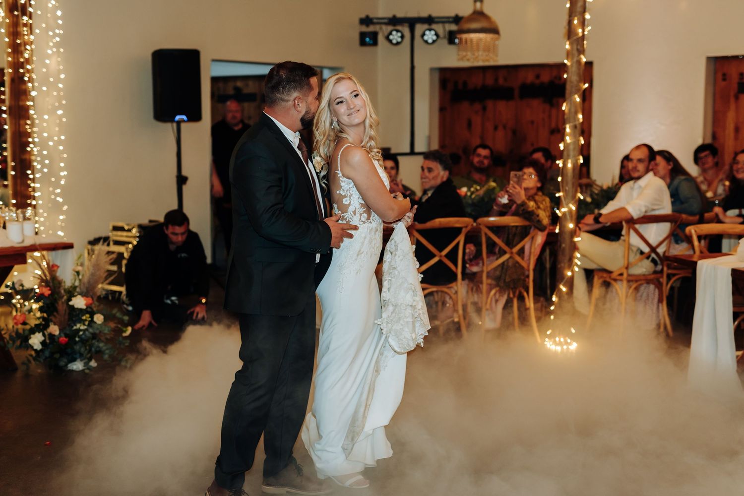 A couple shares their first dance on a misty dance floor illuminated by warm string lights at their wedding reception.