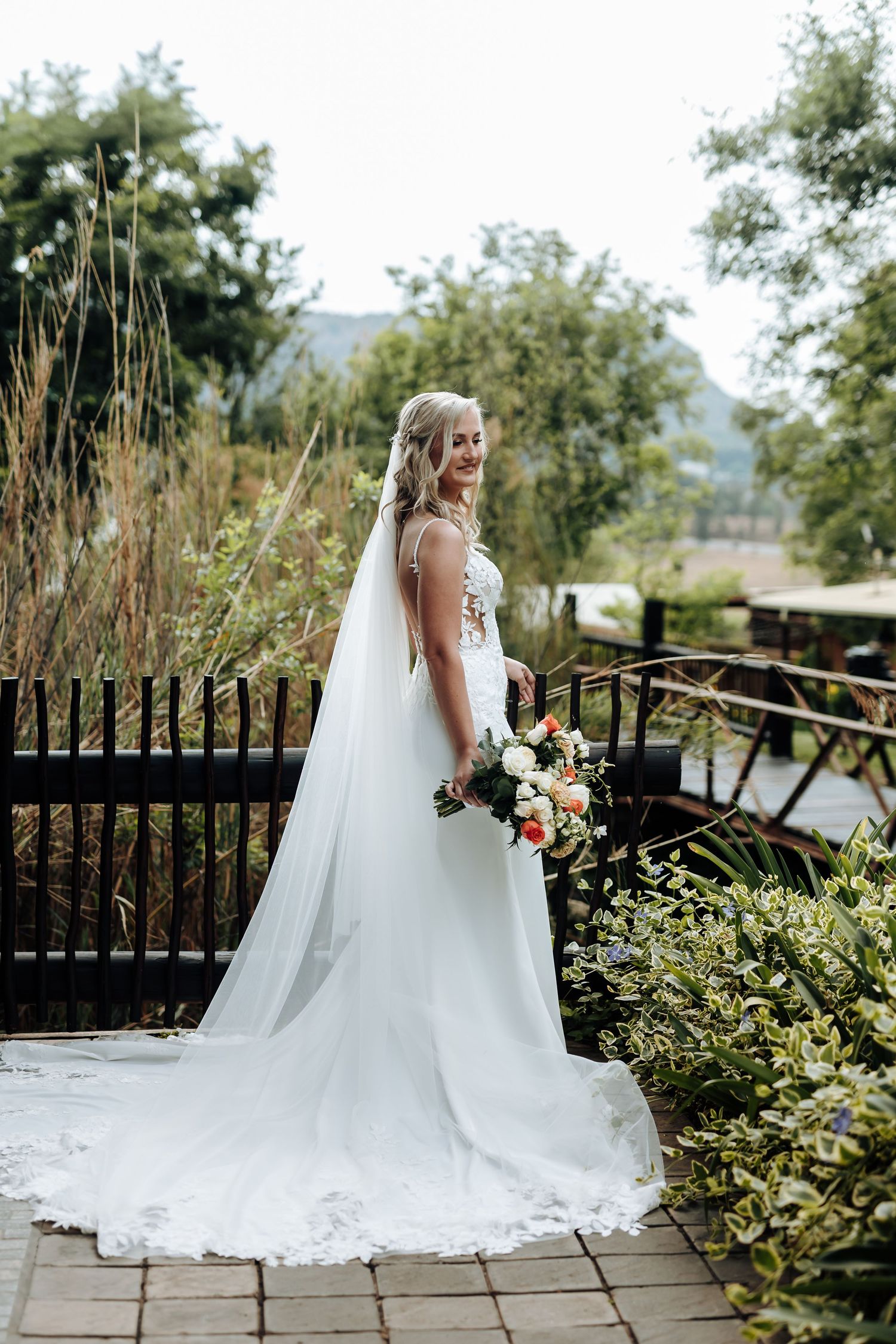 A bride in a flowing white wedding gown and veil poses elegantly on a wooden deck surrounded by lush greenery.