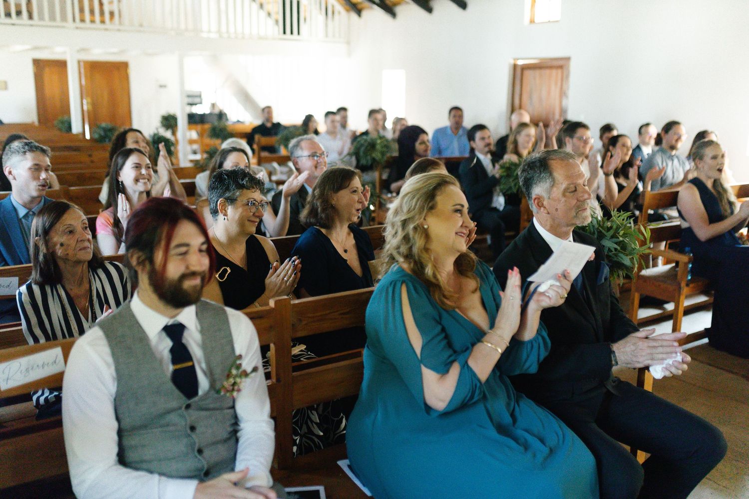 Wedding guests seated in wooden church pews clap and celebrate during a joyful ceremony.