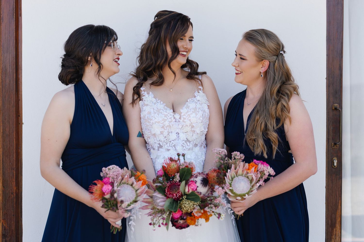 Three women in wedding attire holding colorful bouquets with pink and orange flowers against wooden backdrop.