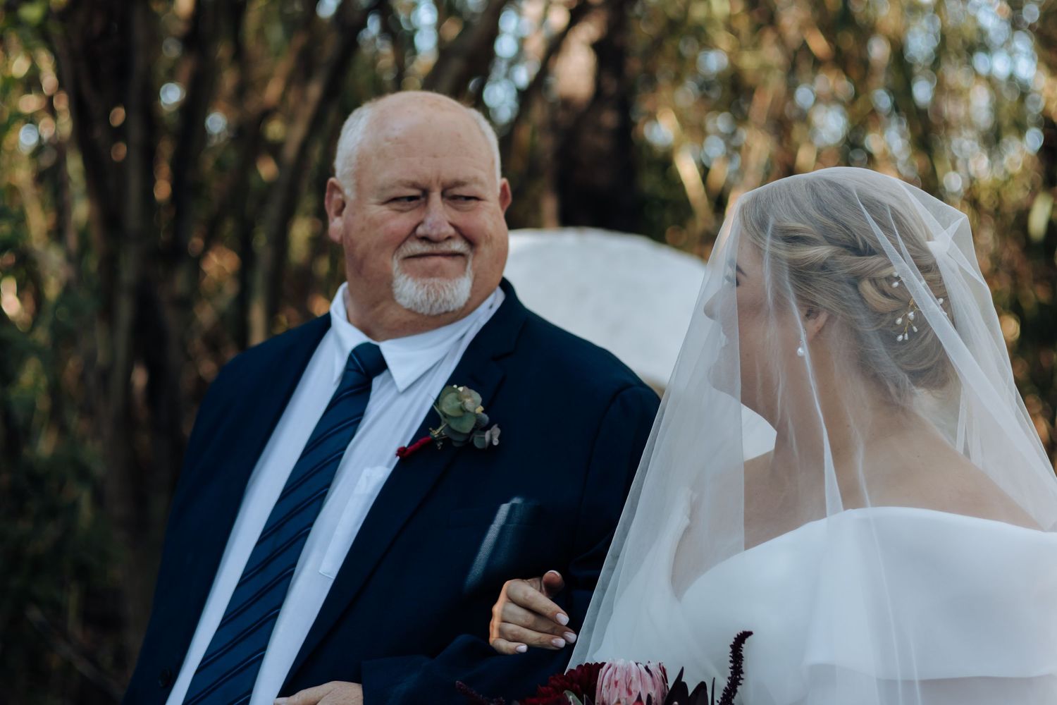 Father of the bride smiling proudly beside his daughter in a white veil at an outdoor wedding ceremony.