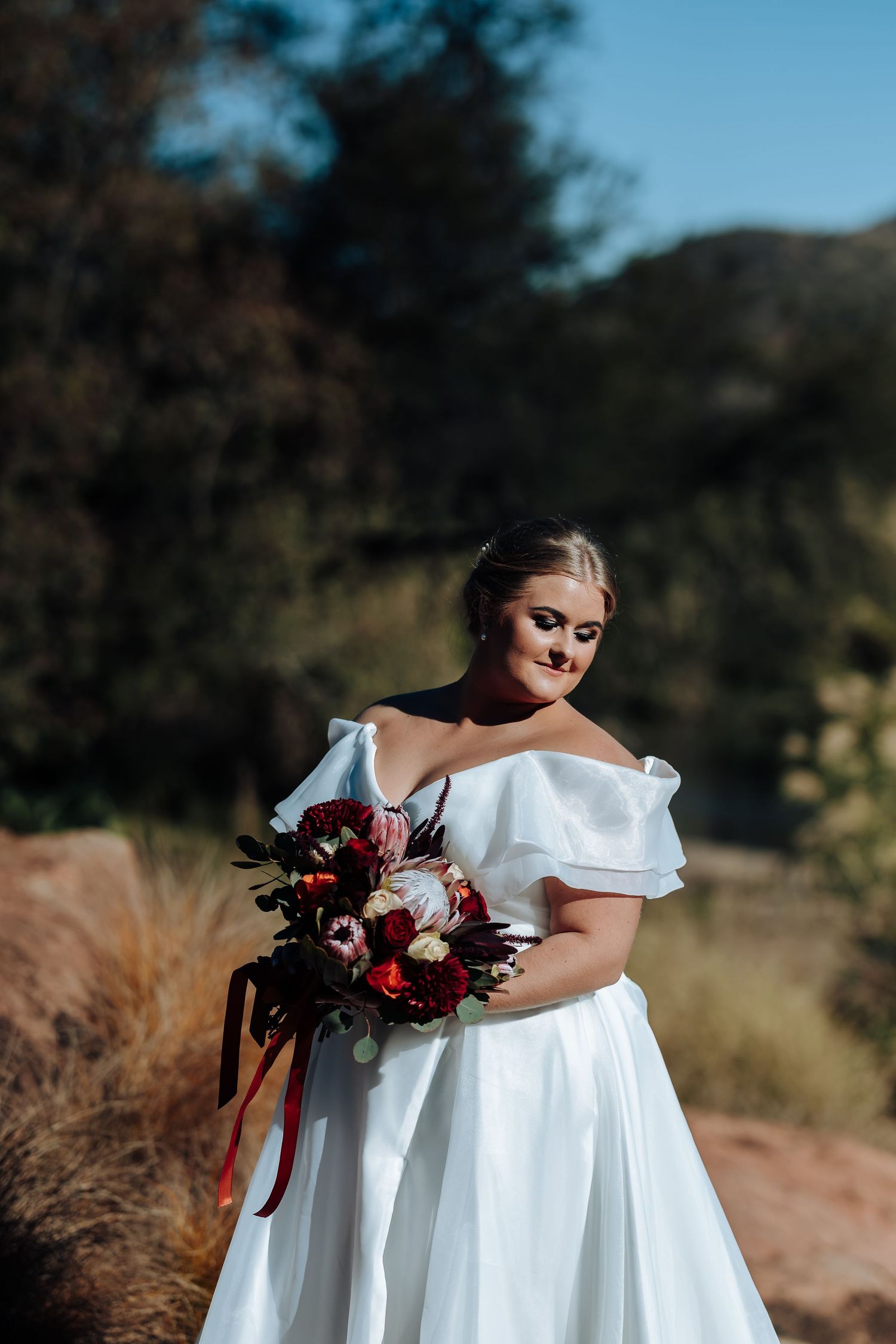 Bride in off-shoulder white gown holding red and white bouquet outdoors in natural setting.