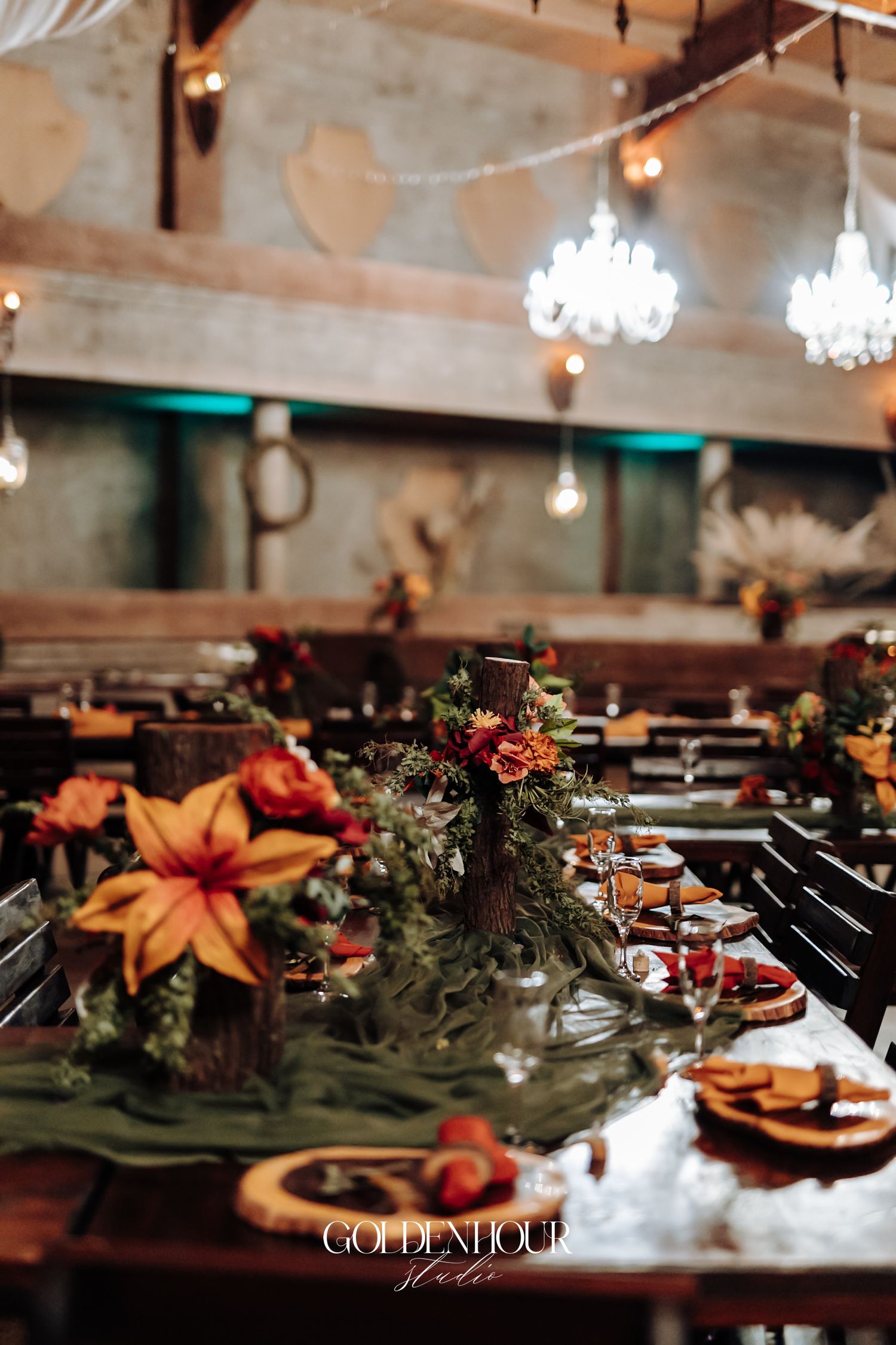 Rustic wedding reception table with autumn floral centerpieces, greenery, and warm Edison bulb lighting in a barn venue.