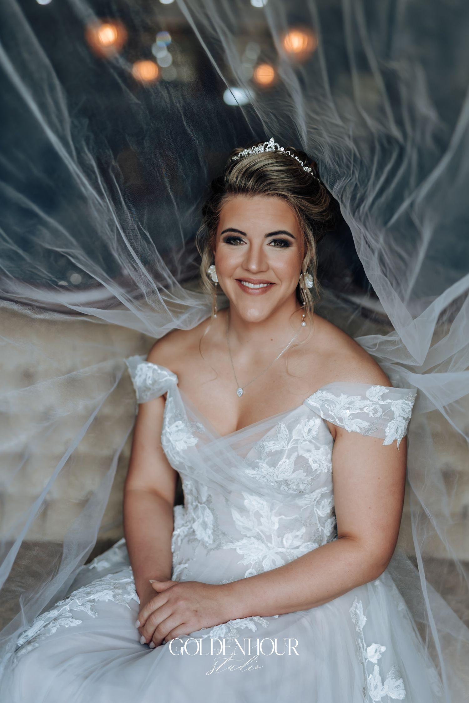 Smiling bride in off-shoulder lace wedding gown with tiara and veil draped elegantly around her.