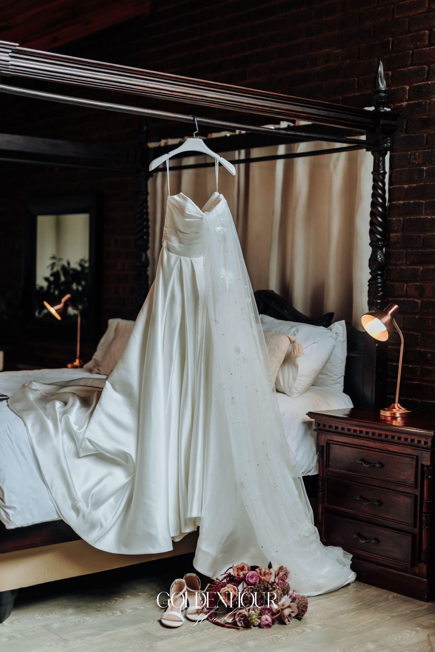 Elegant white satin strapless wedding gown hanging on a four-poster bed in a rustic bridal suite.