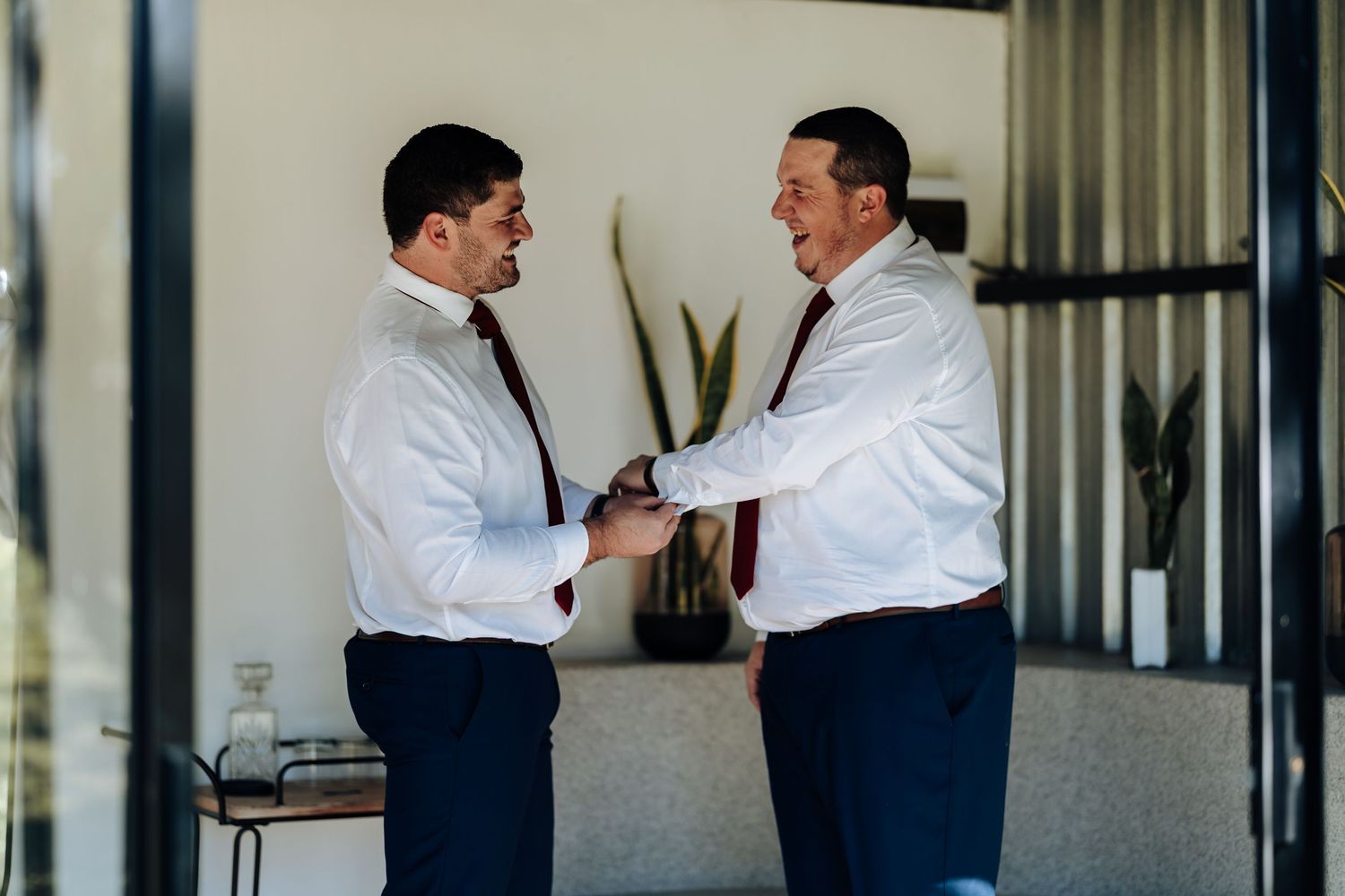 Two groomsmen in white shirts and dark ties help each other prepare for a wedding ceremony indoors.