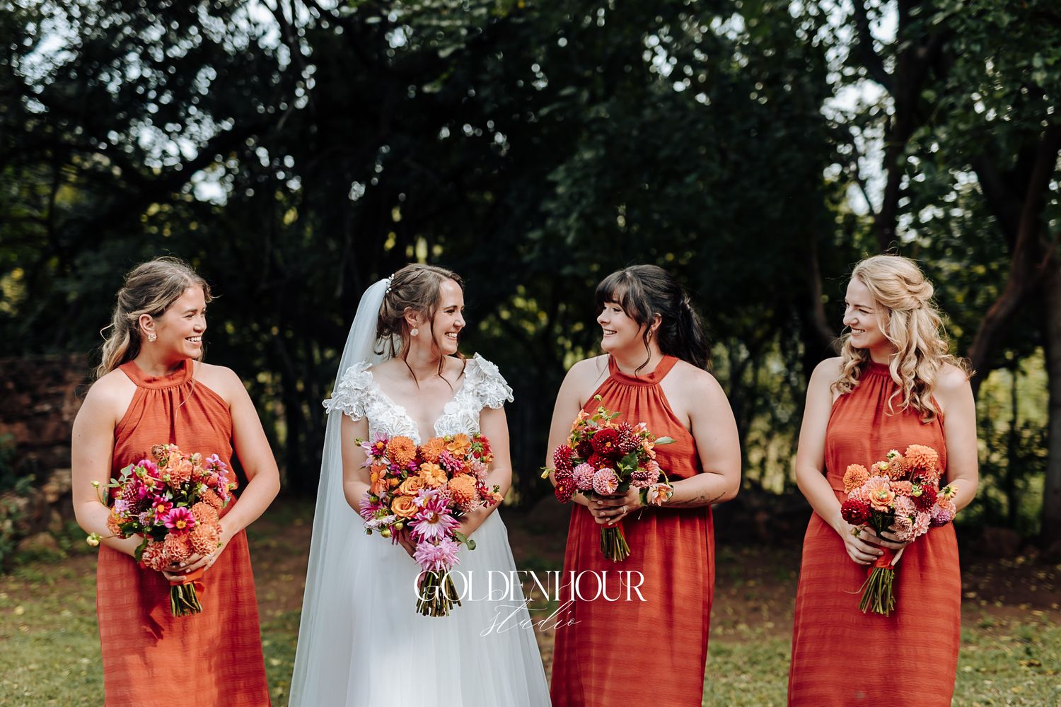 Bride with three bridesmaids in orange dresses holding colorful bouquets outdoors.