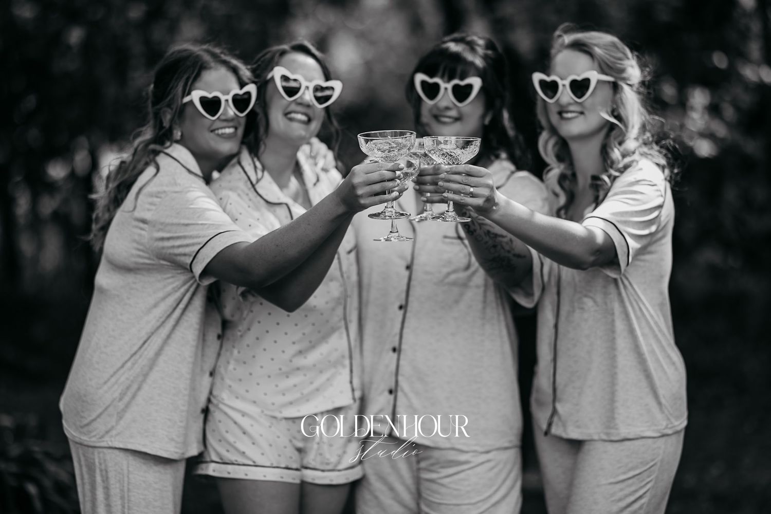 Black and white photo of four bridesmaids in pajamas wearing heart sunglasses toasting with champagne.