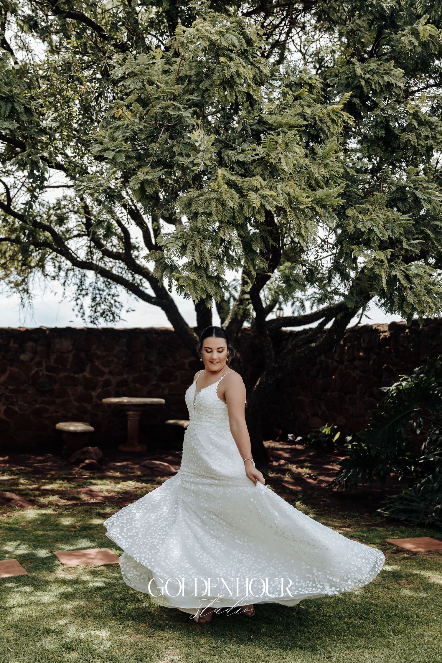 A flowing white wedding dress twirls gracefully against a backdrop of lush green trees in a garden setting.
