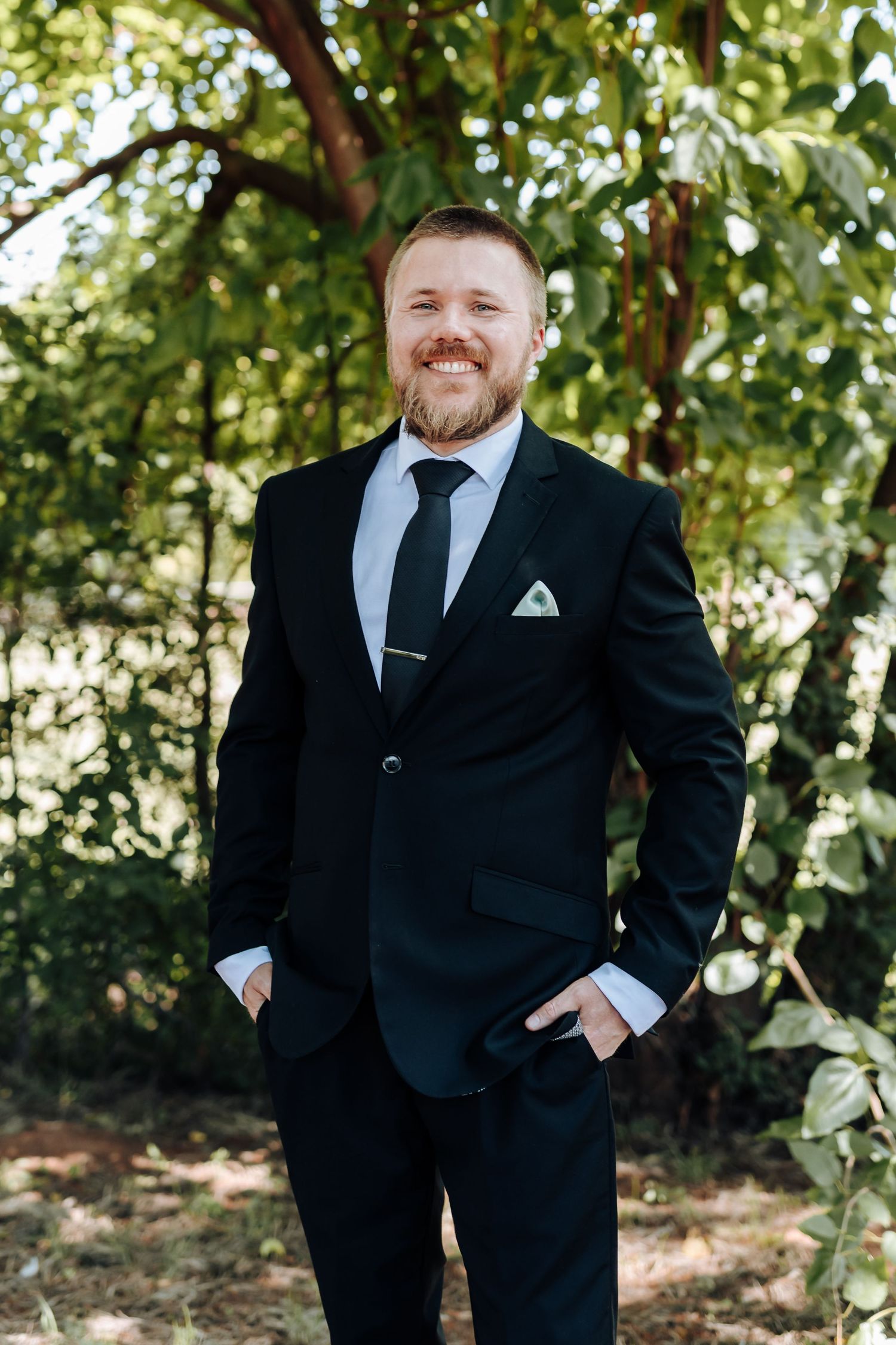 Person in black suit and tie smiling while posing in outdoor garden setting.