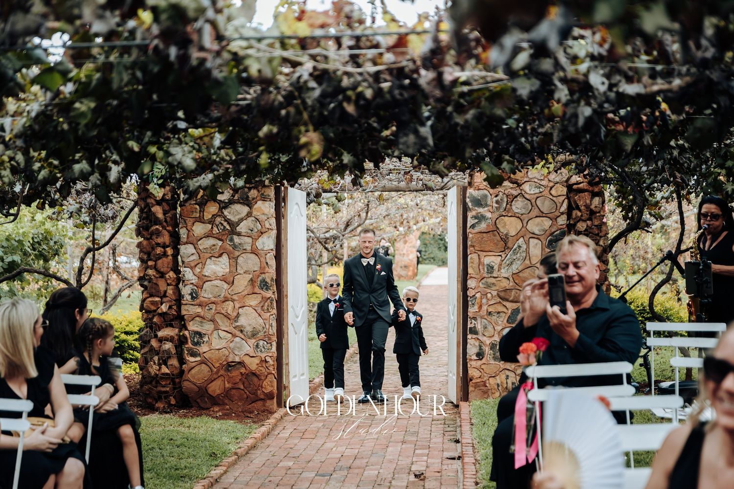 Wedding ceremony guests gather outdoors under trees during a sunny autumn day.