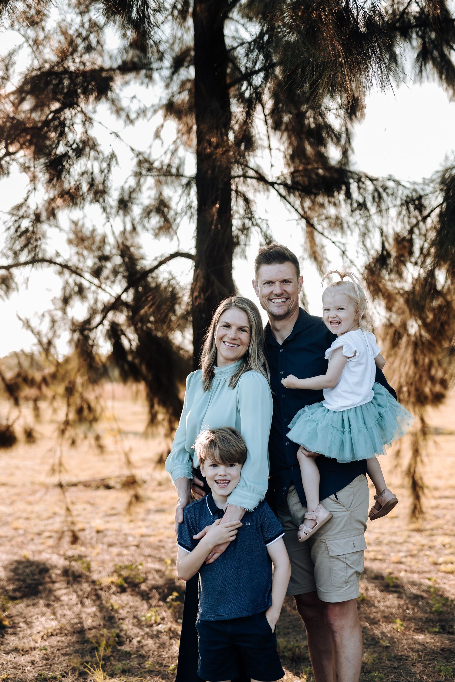 A smiling family of four poses outdoors under a pine tree, with parents holding two young children in a sunny field.