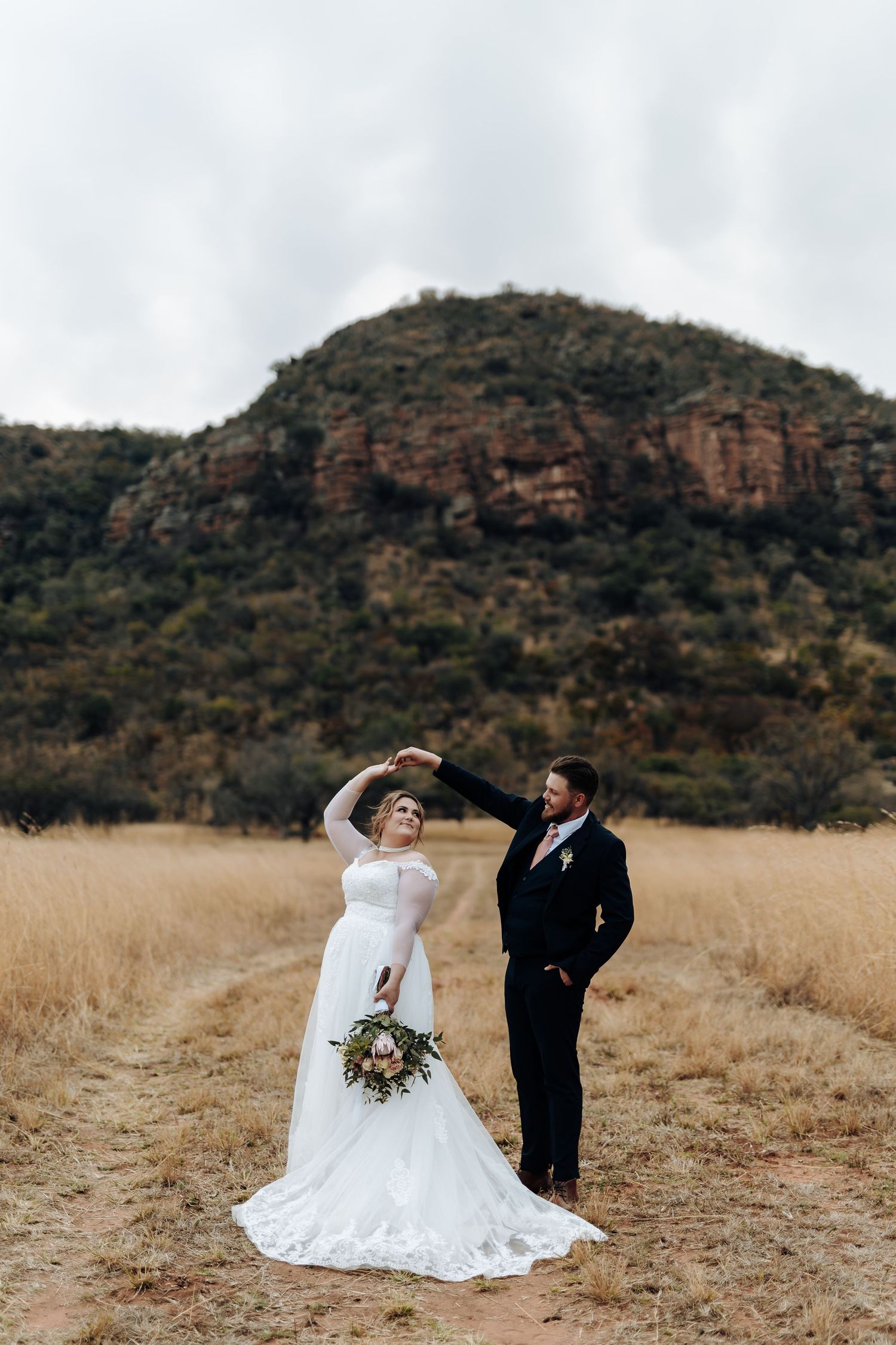 A couple in wedding attire share a romantic moment in a golden field with rugged mountains in the background.