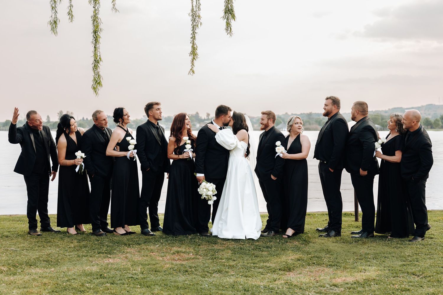 Wedding party in formal black and white attire poses together on grassy hillside overlooking scenic vista.