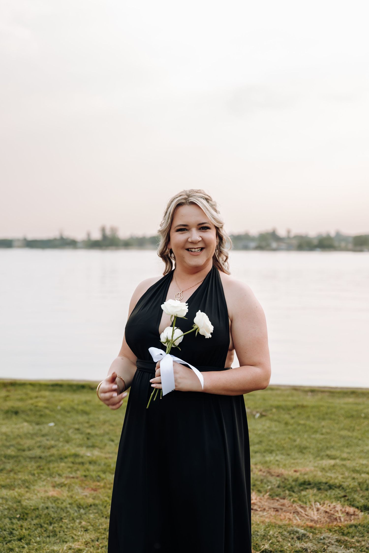 A person in a black formal dress stands by a waterfront holding white flowers on a grassy lawn.