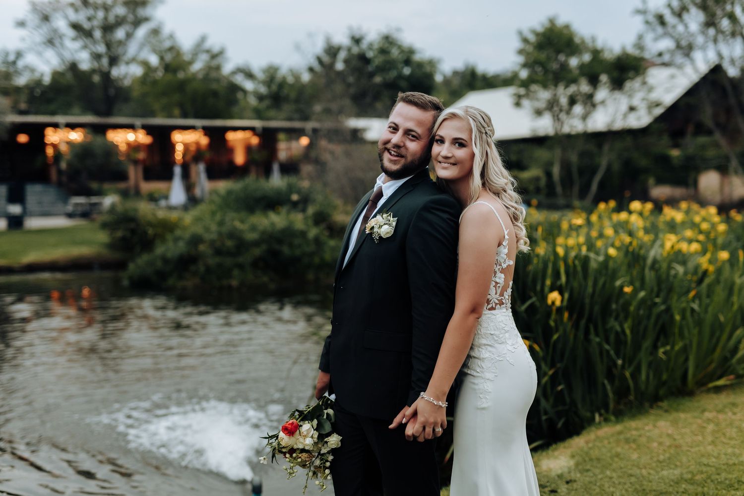 A couple poses together at sunset by a pond with yellow irises and string lights glowing in the background.