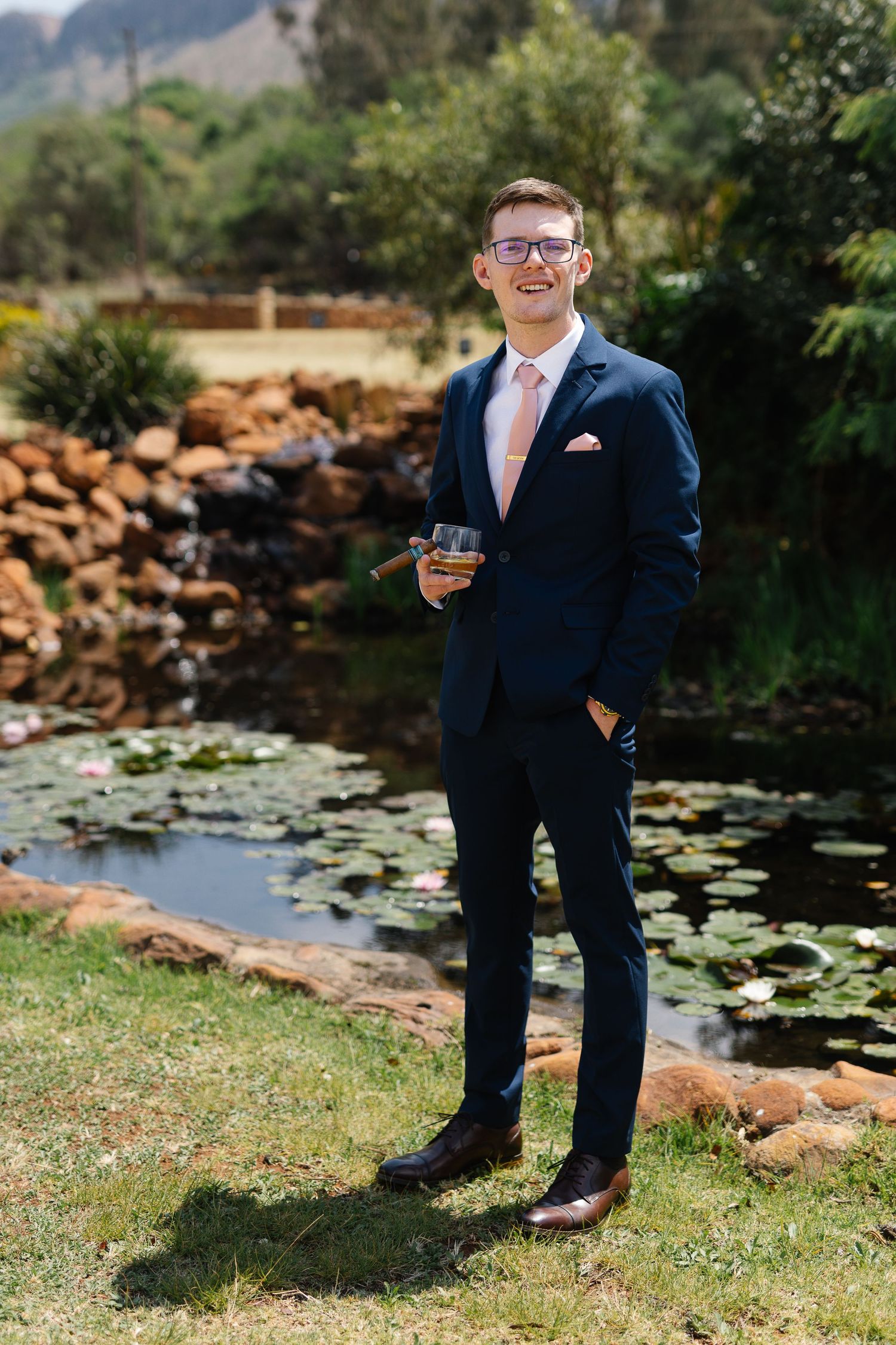A person in a navy suit poses near a tranquil lily pond with rocks and greenery in the background.