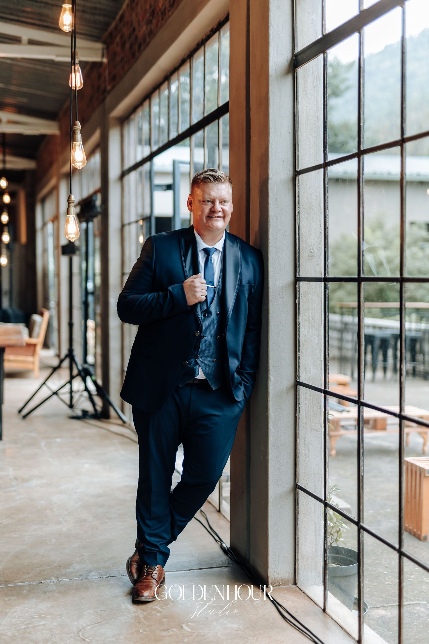 Groom in navy suit leaning against large window frame smiling in elegant industrial venue with Edison bulb lighting.
