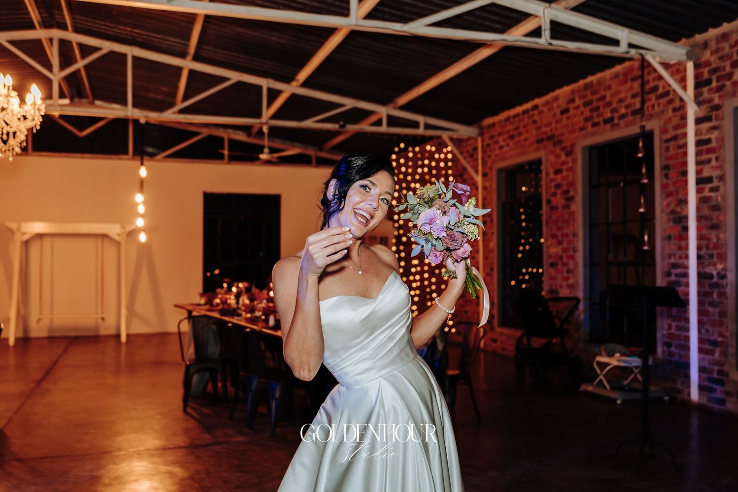 Smiling bride in white strapless gown holding colorful bouquet in warmly lit rustic brick venue.