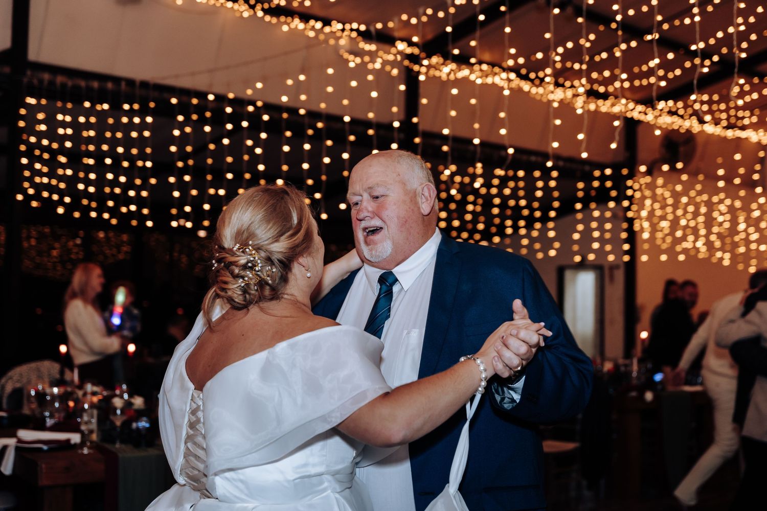Bride and father share emotional father-daughter dance under warm string lights at elegant wedding reception.