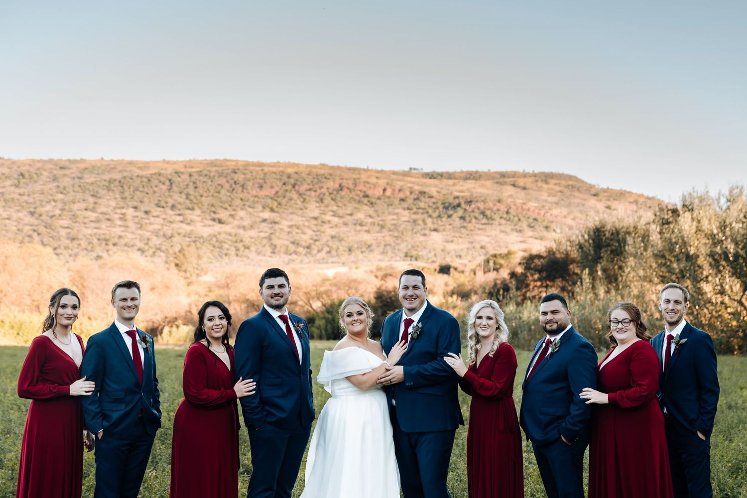 Wedding party in navy suits and burgundy dresses posing outdoors with bride and groom against scenic hill backdrop.