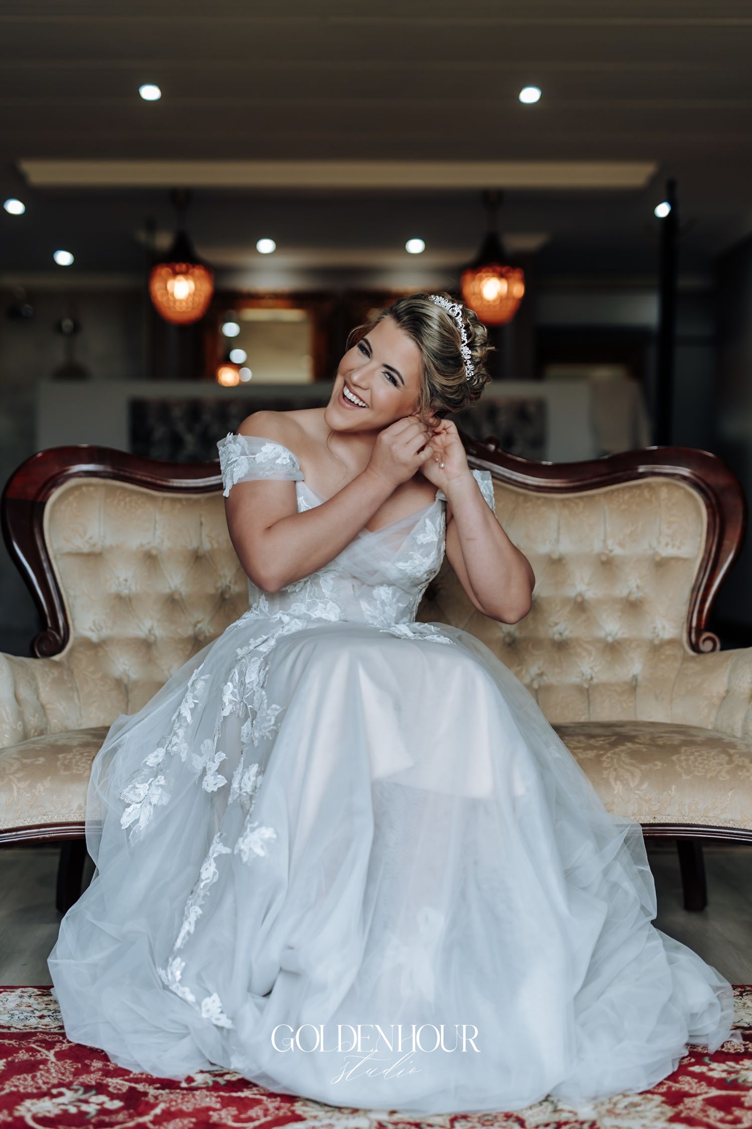 Radiant bride in a floral lace off-shoulder wedding gown sitting on a vintage sofa adjusting her earring.