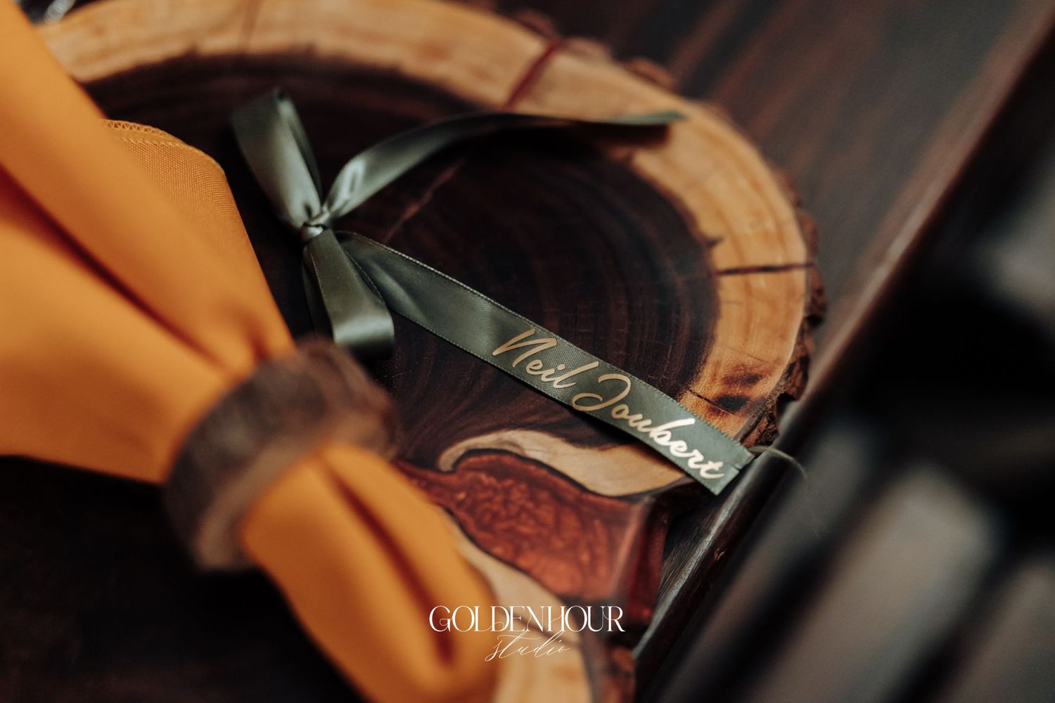 Elegant wedding place card with cursive script tied with black ribbon on rustic wood slice with orange napkin.