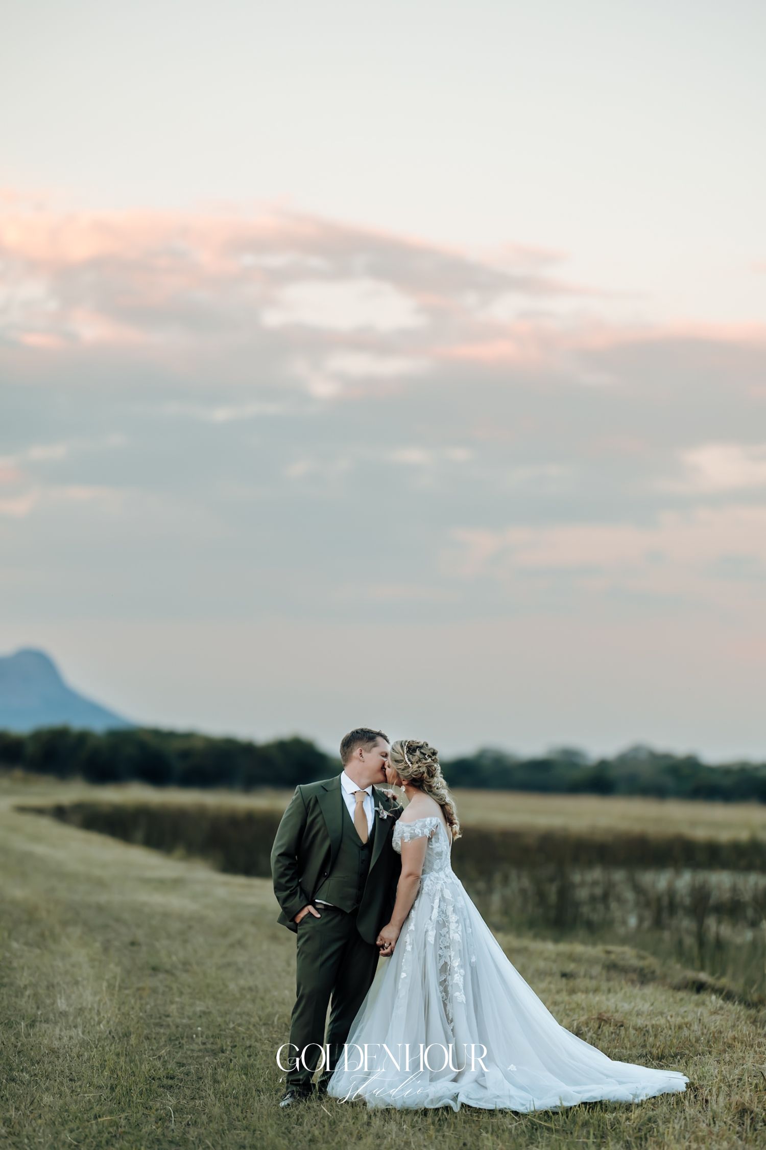Bride and groom sharing a kiss in an open field at dusk with a mountain silhouette in the background.