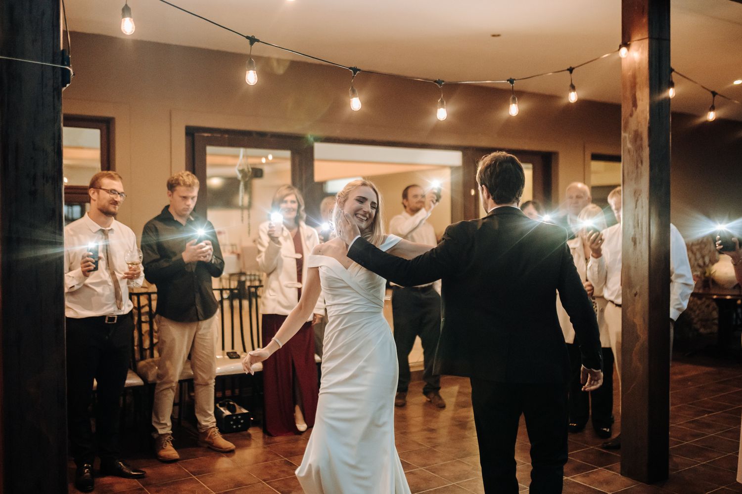 Dancing couple shares their first dance surrounded by wedding guests under warm indoor lighting.