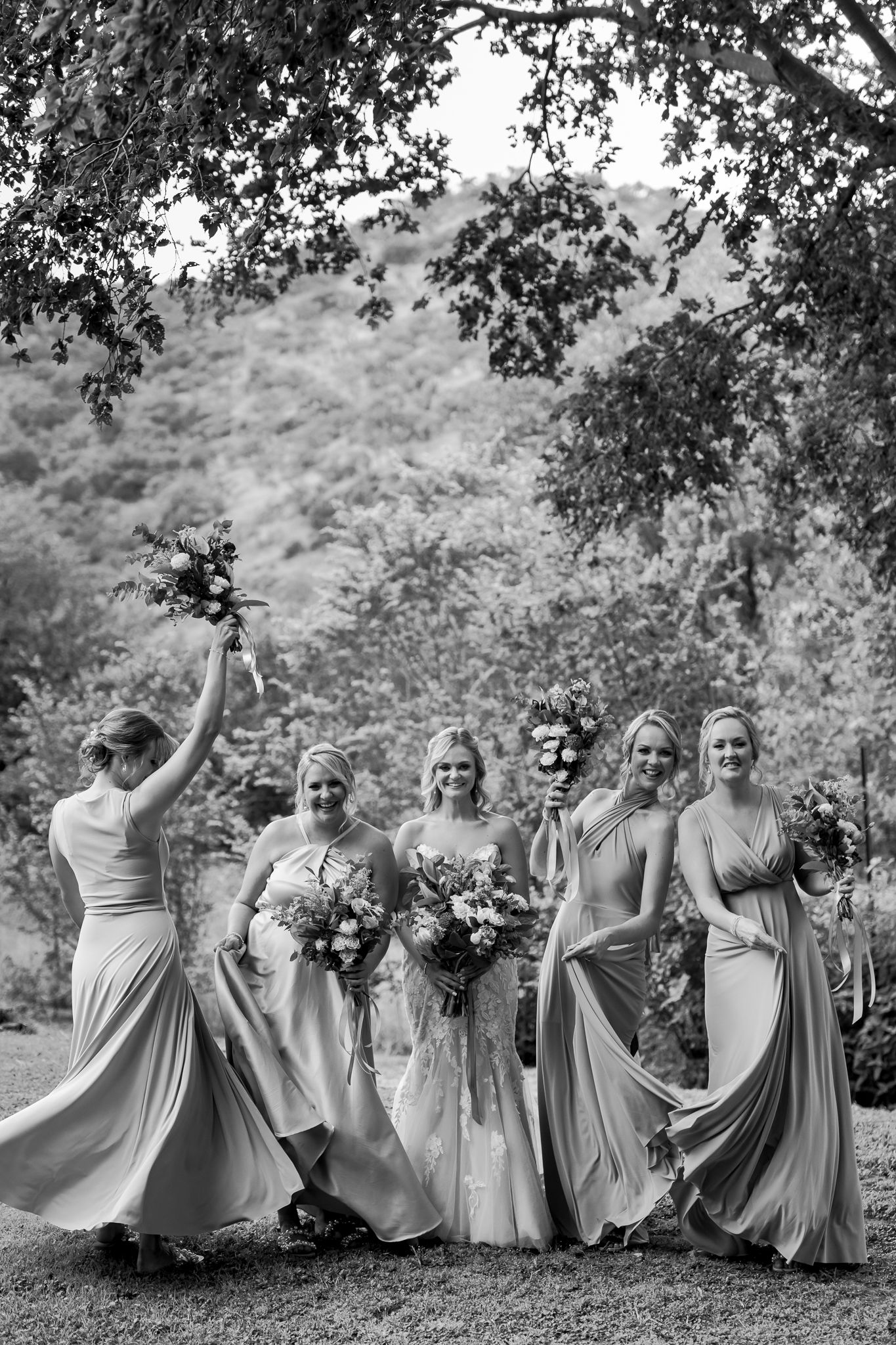A joyful wedding party in flowing dresses twirls and dances together outdoors with mountains in the background.