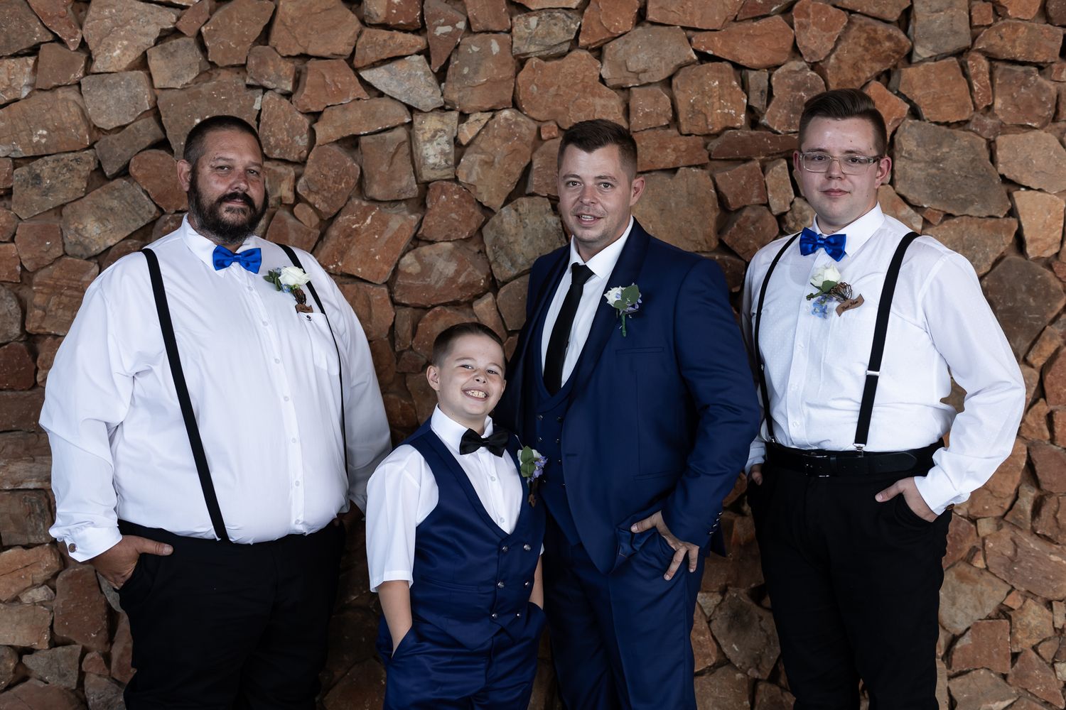 Wedding party members in blue and white formal attire pose against a rustic stone wall.