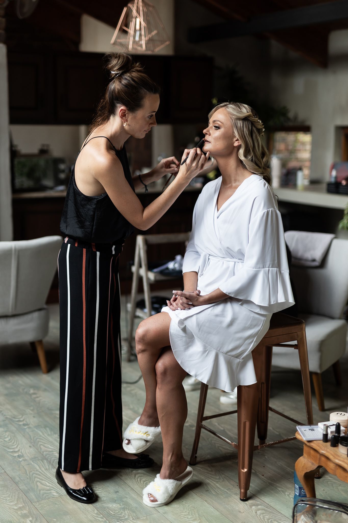 A makeup artist applies cosmetics to a woman in a white robe during wedding preparations.