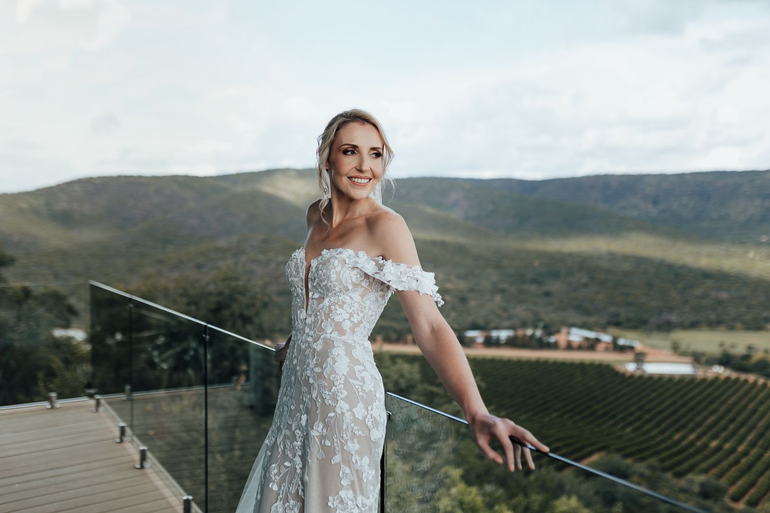 Bride in detailed wedding gown stands against scenic vineyard and mountain backdrop.