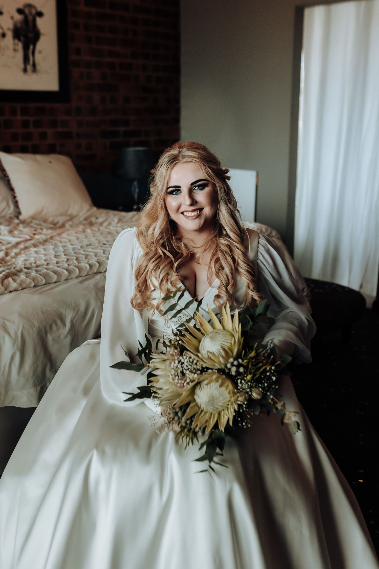 Bride sitting on bed holding dried flower bouquet in front of exposed brick wall.
