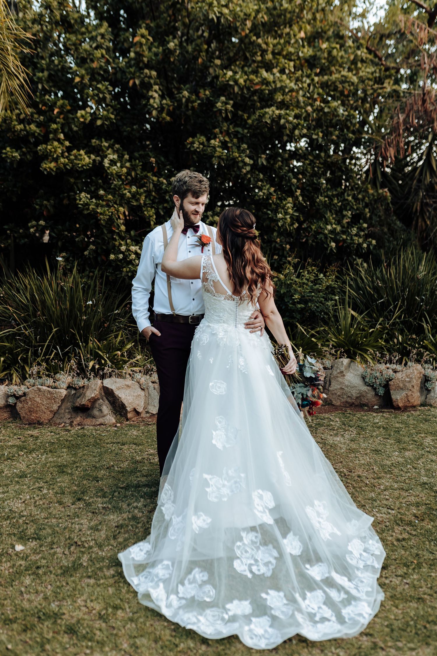 Wedding couple in formal attire standing in garden with bride wearing floral lace gown.