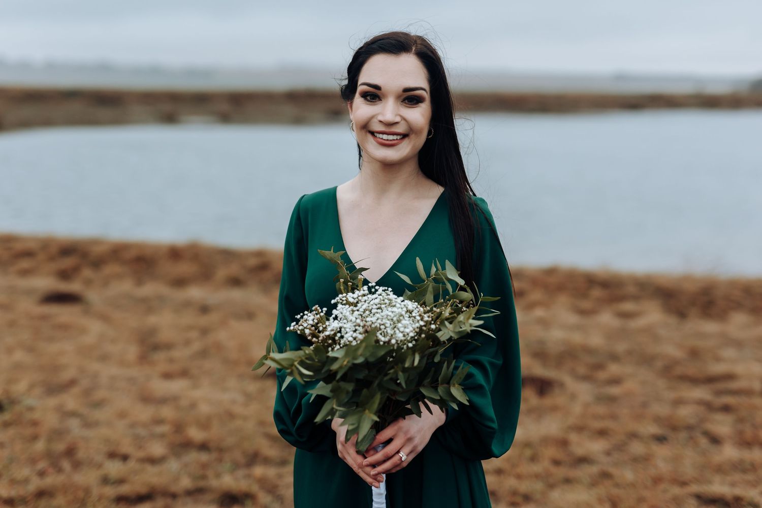 Bridesmaid poses with bouquet by waterfront in emerald green dress.