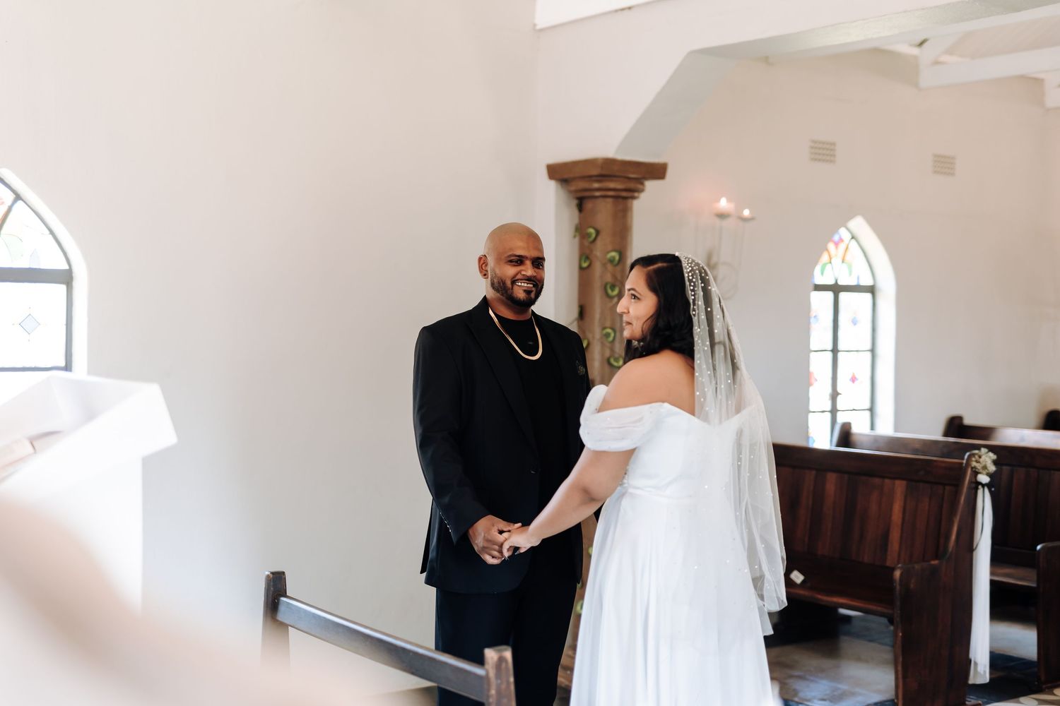 A couple sharing their wedding vows in a bright white chapel with elegant chandeliers.