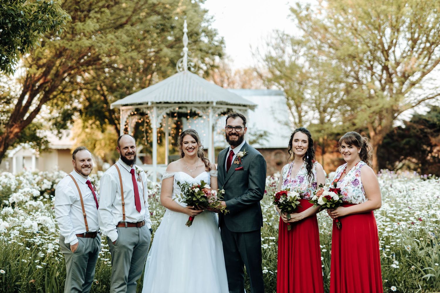 Full wedding party poses outdoors with bridesmaids in red and groomsmen in suspenders.