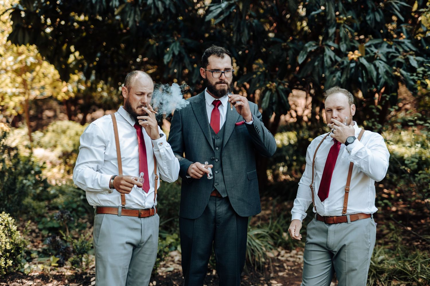Three men in wedding attire drinking mini Coca-Cola bottles against foliage background.