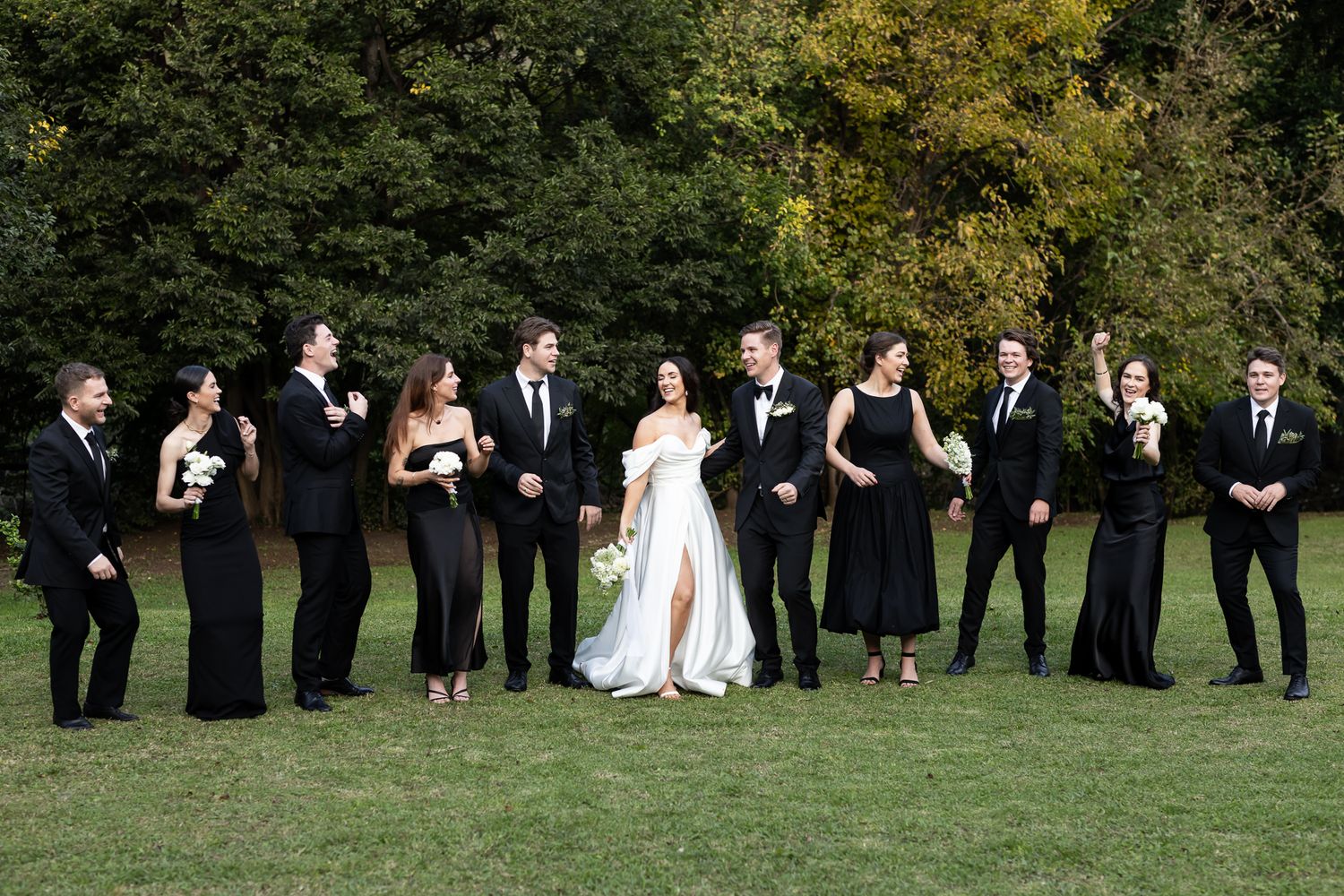 A formal wedding party in black attire poses on grass with autumn trees in the background.