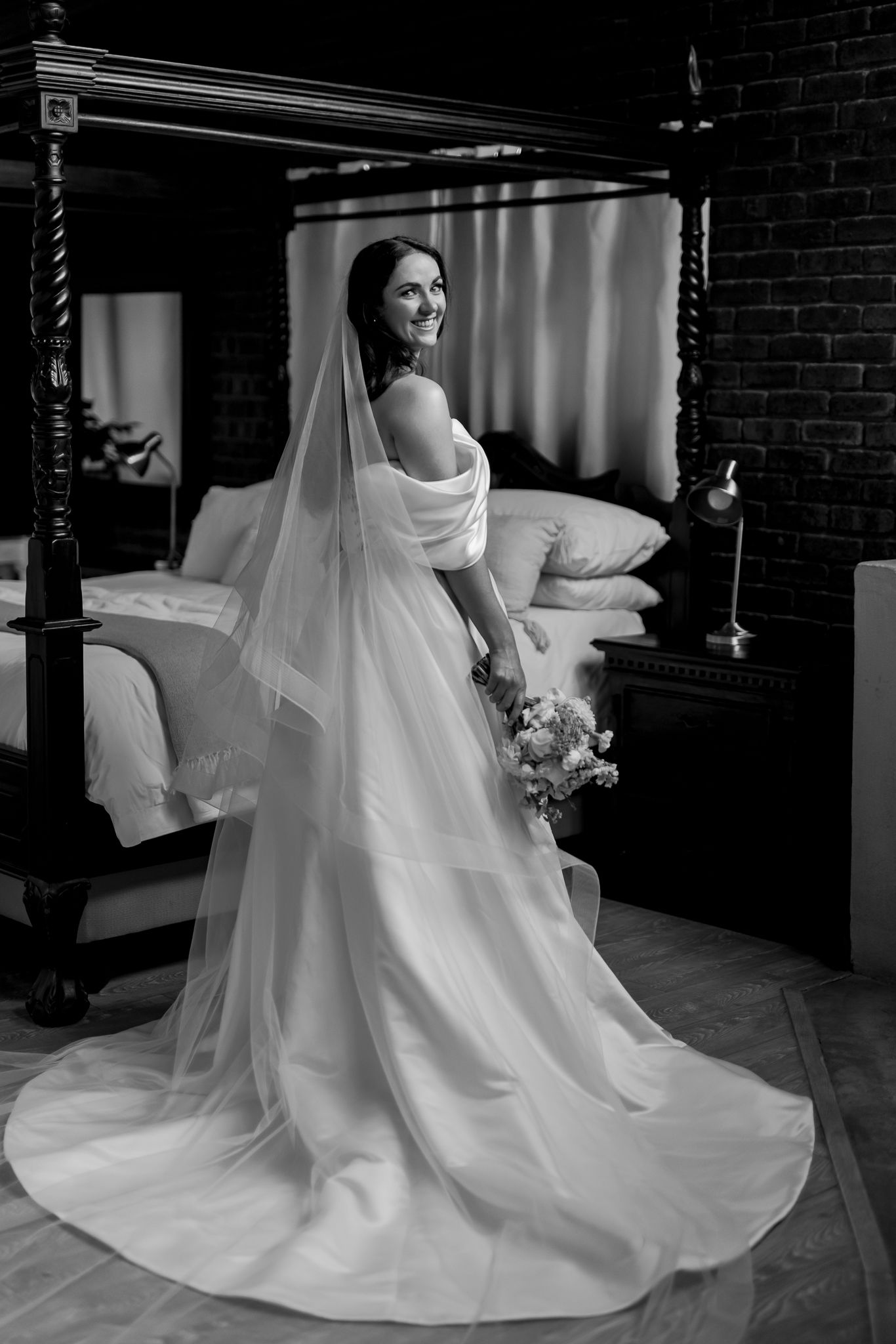 Classic black and white portrait of bride in flowing dress and veil by canopy bed.