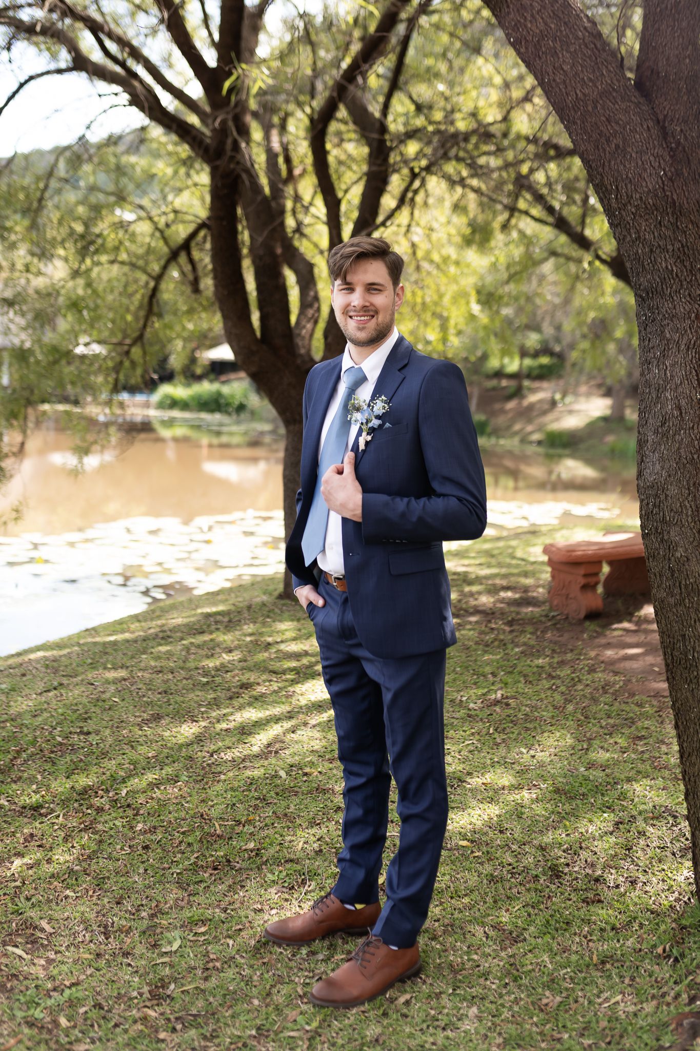 A groom in a navy suit poses by a lakeside with trees in the background.