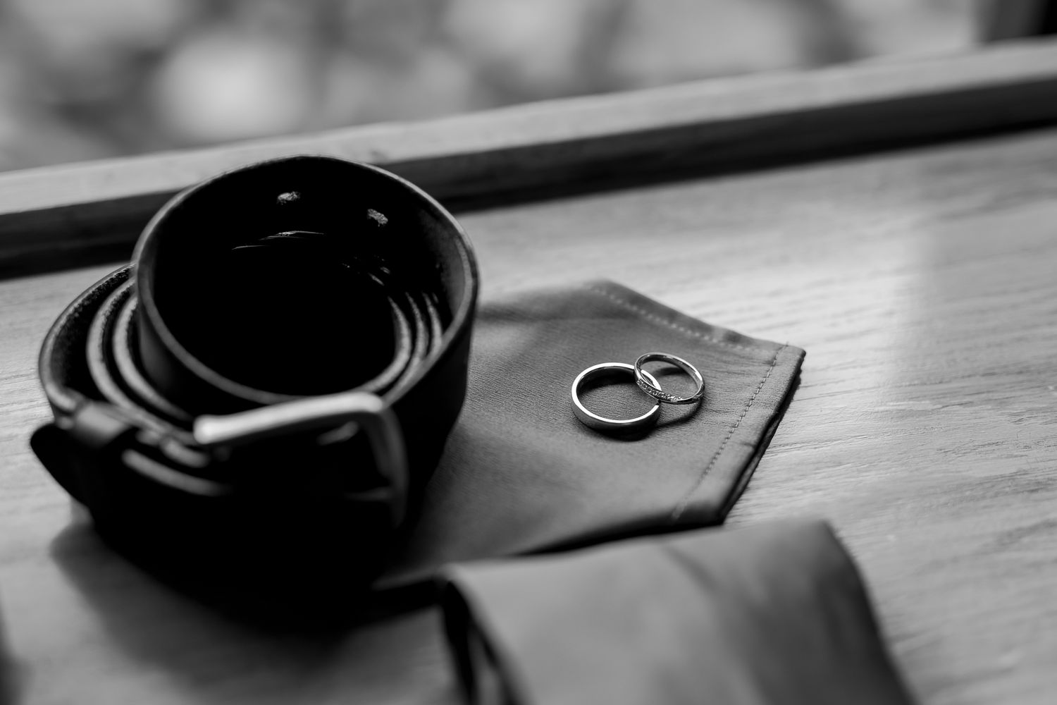 Black leather belt and wedding rings arranged on wooden surface in black and white.