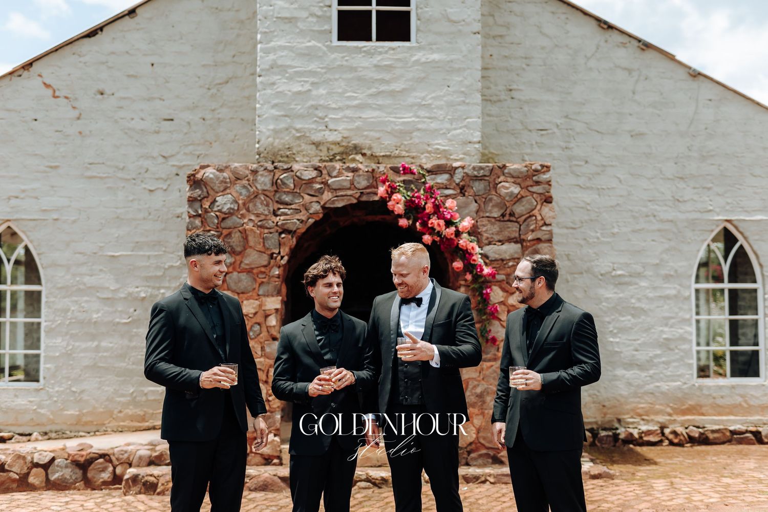 Groomsmen in black suits stand together laughing and looking at their phones against a white stone wall.