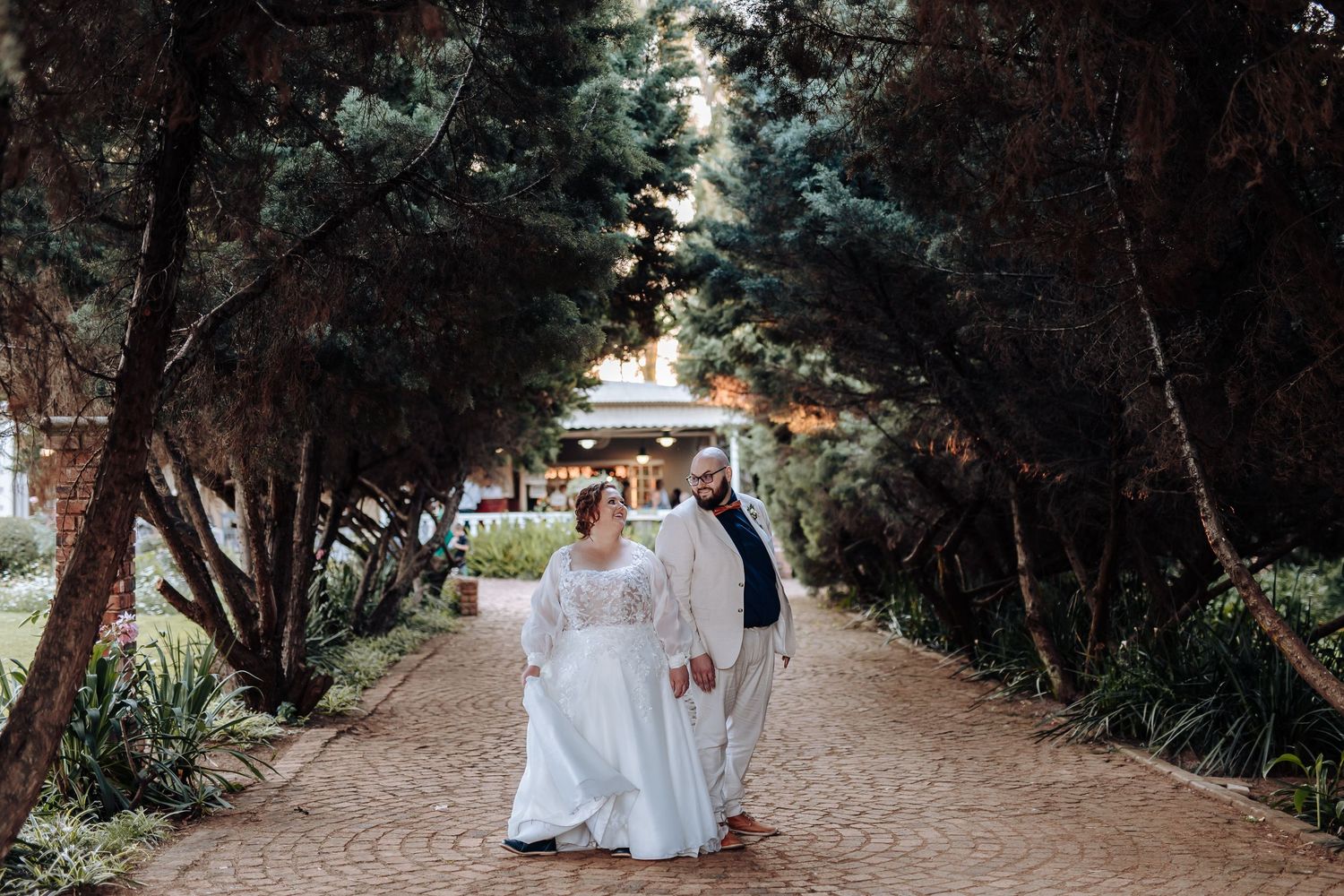 A couple in wedding attire walks down a tree-lined path at a rustic outdoor venue.