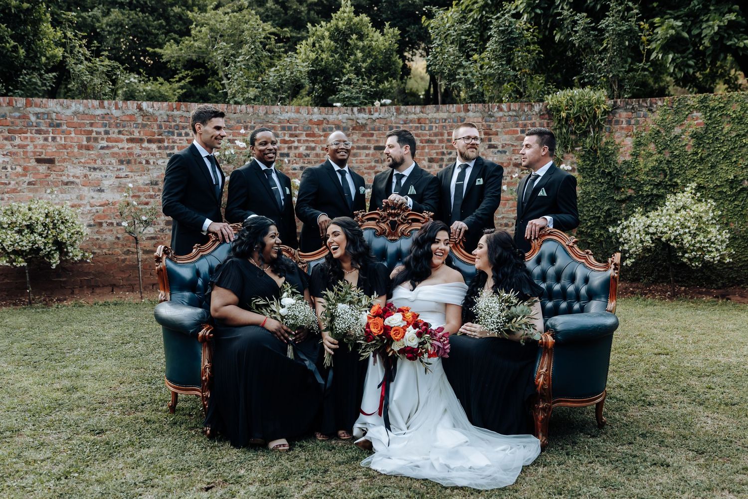 Wedding party relaxes on grass for casual group photos against brick wall backdrop.