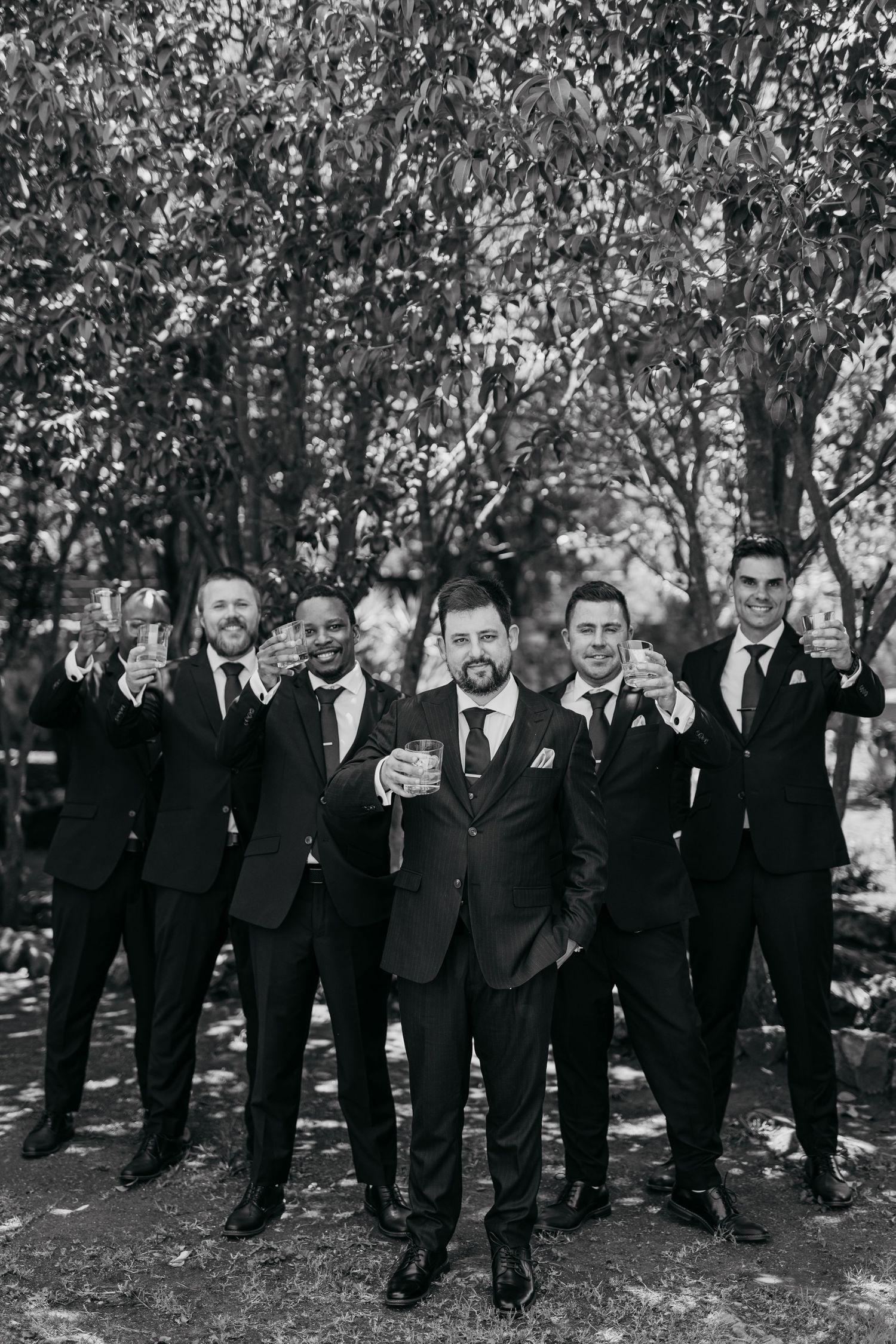 Large group of groomsmen in black suits posing together outdoors.