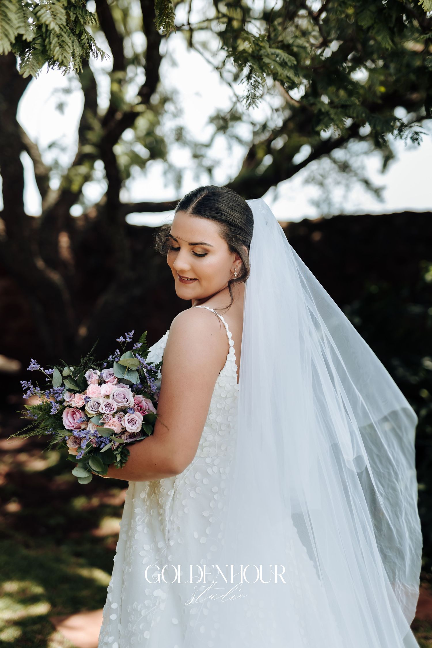 A bride in a white lace dress holds a pink and purple bouquet against a natural outdoor backdrop.