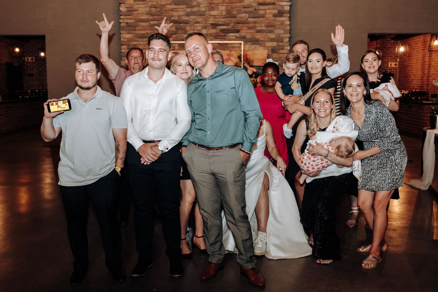 Large group celebrates at wedding reception with energetic poses in front of stone wall.